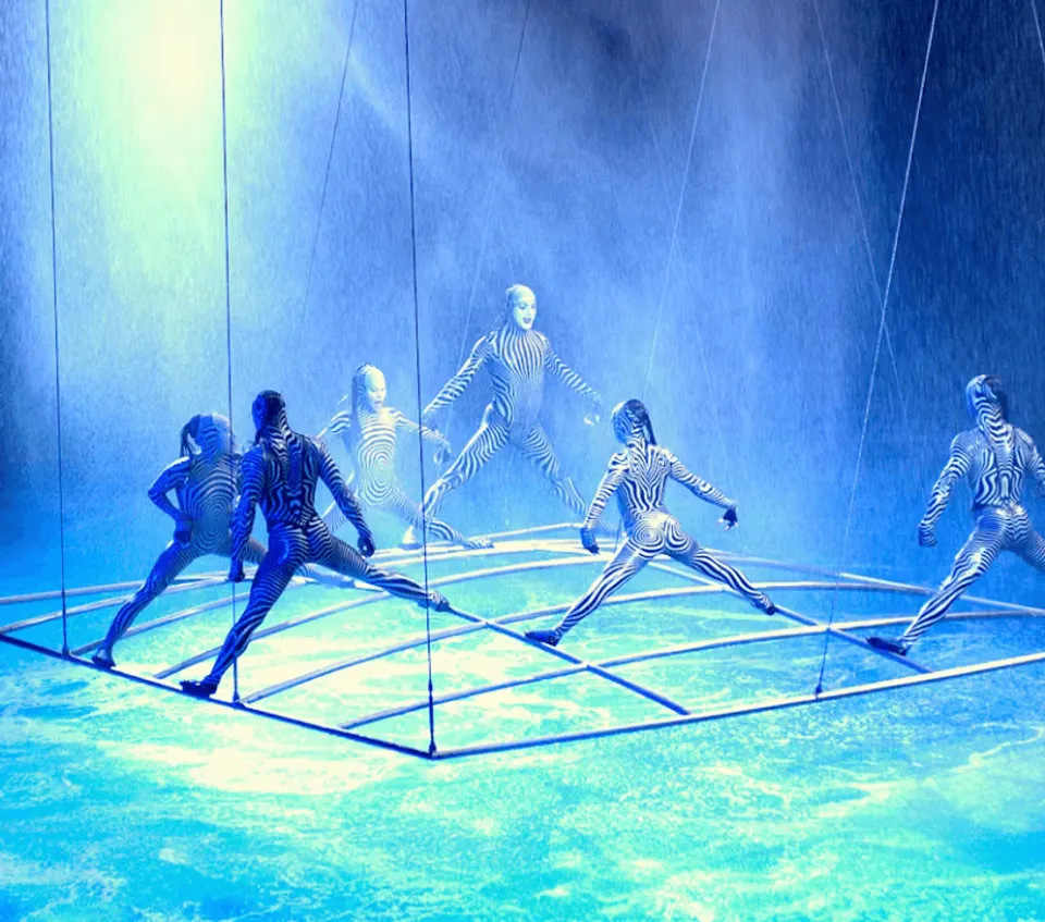 O acrobats performing on a suspended frame above blue-lit water